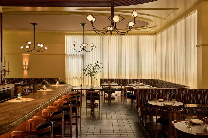 A modern restaurant interior with a long bar, wooden chairs, round tables, patterned bench seating, tiled floor, and warm lighting from decorative ceiling fixtures.