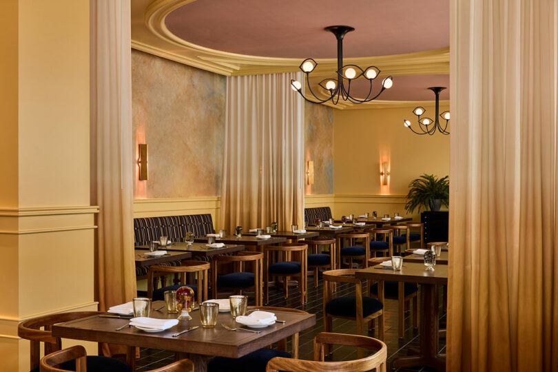 A spacious restaurant dining area with wooden tables, upholstered chairs, soft lighting, wall sconces, and chandeliers, featuring neutral tones and elegant decor.