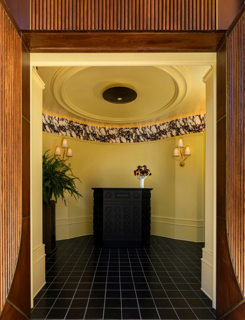 A small entryway with yellow walls, black tiled floor, a black ornate console table, decorative wallpaper border, wall sconces, plants, and a ceiling light.
