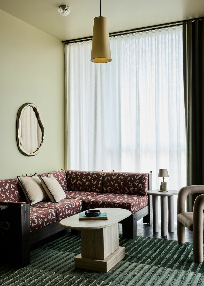 A living room with a patterned red sectional sofa, two beige chairs, a wooden coffee table, a side table with a lamp, a mirror, and large sheer curtains letting in natural light.