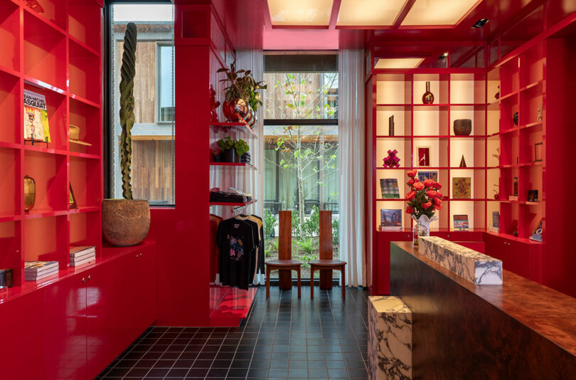 A modern retail space featuring glossy red shelves with books, decor, and apparel, a marble counter, two wooden chairs, and large windows overlooking a small garden.