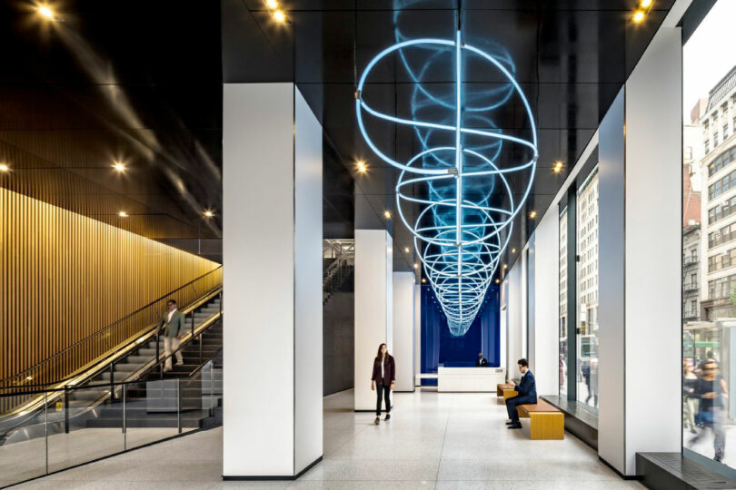Modern building lobby with tall windows, a blue spiral light fixture on the ceiling, seating benches, columns, and people walking or sitting; escalator visible on the left.