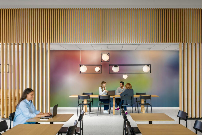 Four people are seated at tables in a modern, brightly lit room with wooden slats, colorful wall art, and contemporary hanging lights; three are conversing, one works on a laptop.