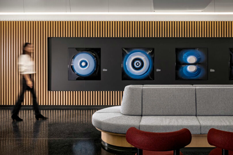 A blurred person walks past abstract circular artwork displayed on a wood-paneled wall in a modern lobby with gray and red seating.