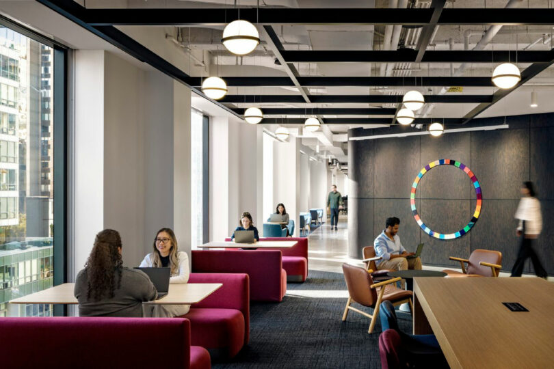 Modern office lounge with people working at tables and using laptops, large windows with city views, and a colorful circular wall art in the background.