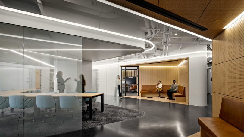 Modern office interior with glass walls, a conference table, and people sitting and standing in a lounge area under bright ceiling lights.