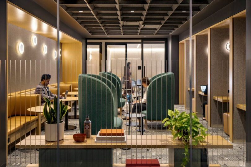 Modern office lounge with green high-back chairs, people working at tables, warm lighting, plants, and books on a glass divider in the foreground.