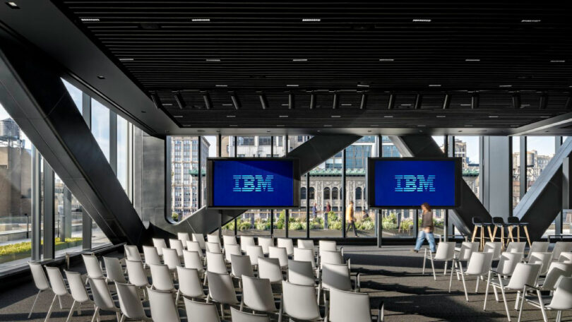 Rows of empty white chairs face two large screens displaying the IBM logo in a modern, glass-walled conference room with city views.