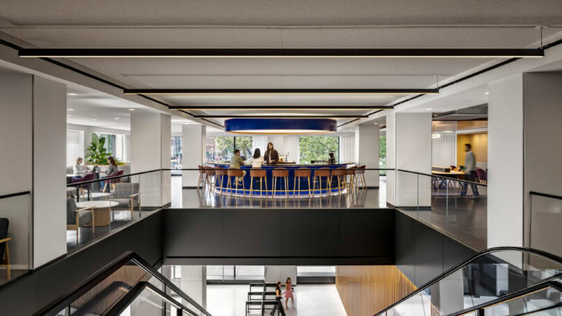 Modern office interior with a central bar-height table where people are seated, surrounded by glass railings and adjacent workspaces, viewed from an upper floor near an escalator.