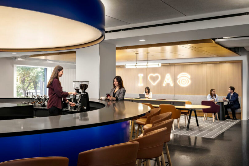 A modern office lounge with people talking and working; one person makes coffee at a counter, others sit at tables. A neon sign on the wall reads "I ?? A" with a wifi symbol.