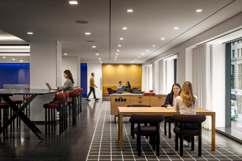 Modern office space with several people working at tables and counters, large windows, and bright ceiling lights.