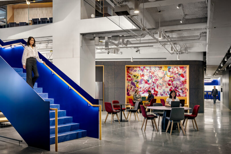 Modern office interior with people working at tables, a woman descending bright blue stairs, and a colorful abstract artwork on the wall.