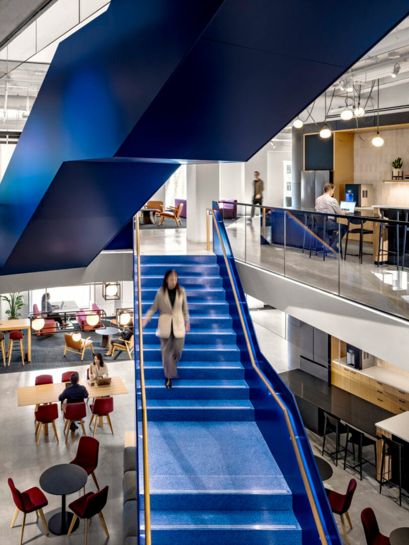 A modern office interior with a bright blue staircase, people working at tables and counters, and contemporary furniture throughout the open space.