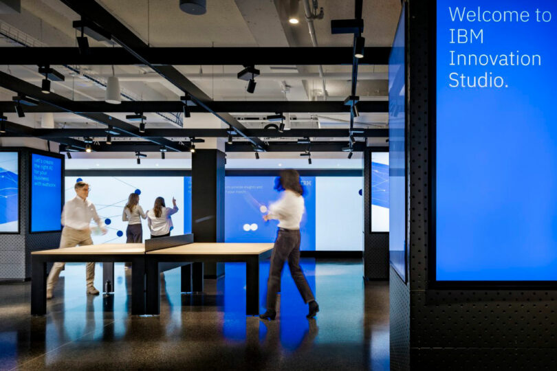 Four people interact with digital displays in a modern workspace with a sign reading "Welcome to IBM Innovation Studio" on a blue screen.