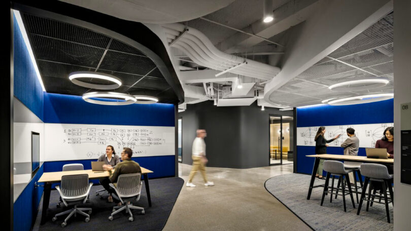 Modern office space with groups of people collaborating at tables and whiteboards, featuring circular ceiling lights and exposed ductwork.