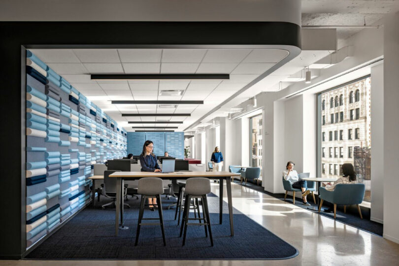 Modern office space with large windows, several people working at desks and tables, and blue-accented wall panels. Natural light fills the room.