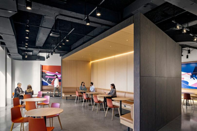 Modern office break area with round tables and orange chairs; several people are sitting, talking, or working, with abstract art on the walls and exposed ceiling features.