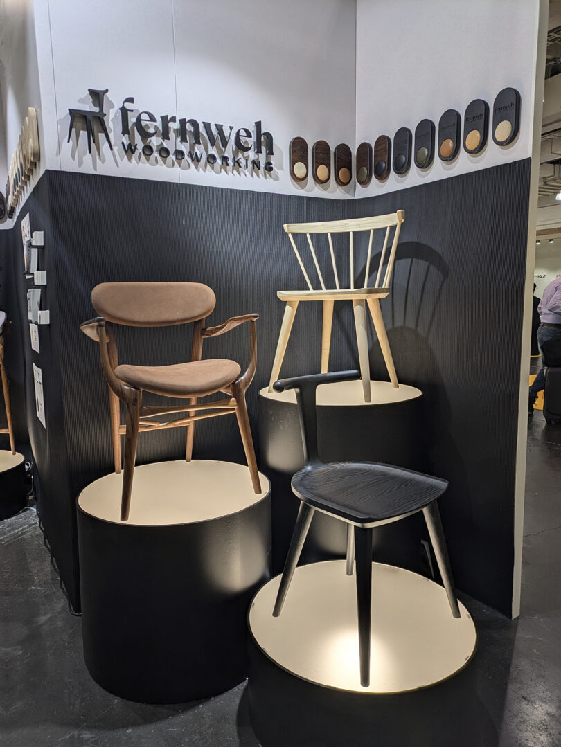 Three wooden chairs are displayed on round pedestals at a Fernweh Woodworking exhibit, with wood finish samples mounted on the wall above.