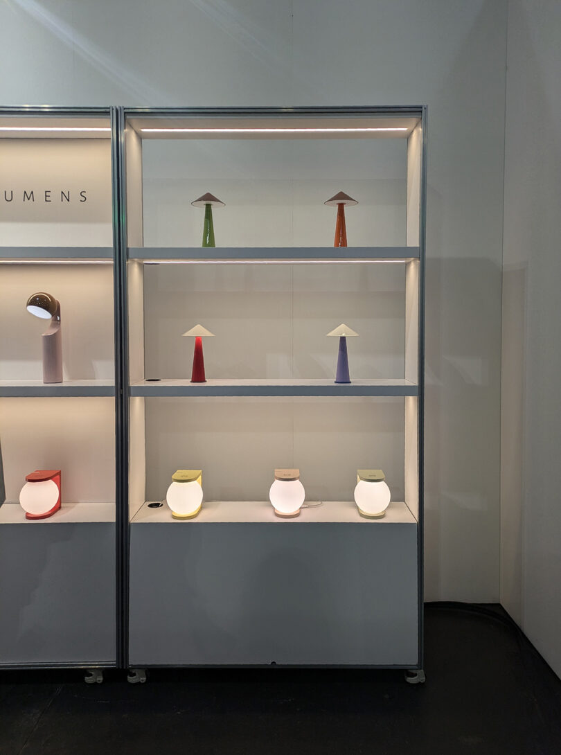A display shelf with various modern table lamps in different colors and shapes, illuminated and arranged on three shelves against a white background.