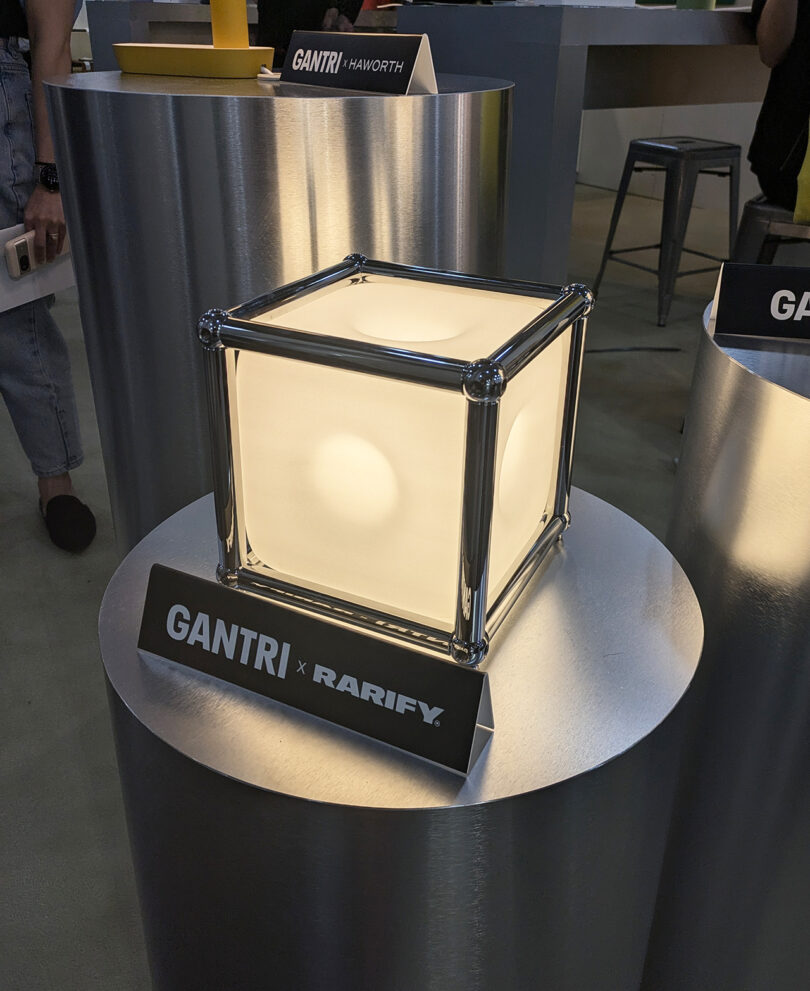 A cube-shaped lamp with metal frame and frosted panels sits on a metallic pedestal labeled "GANTRI x RARIFY.