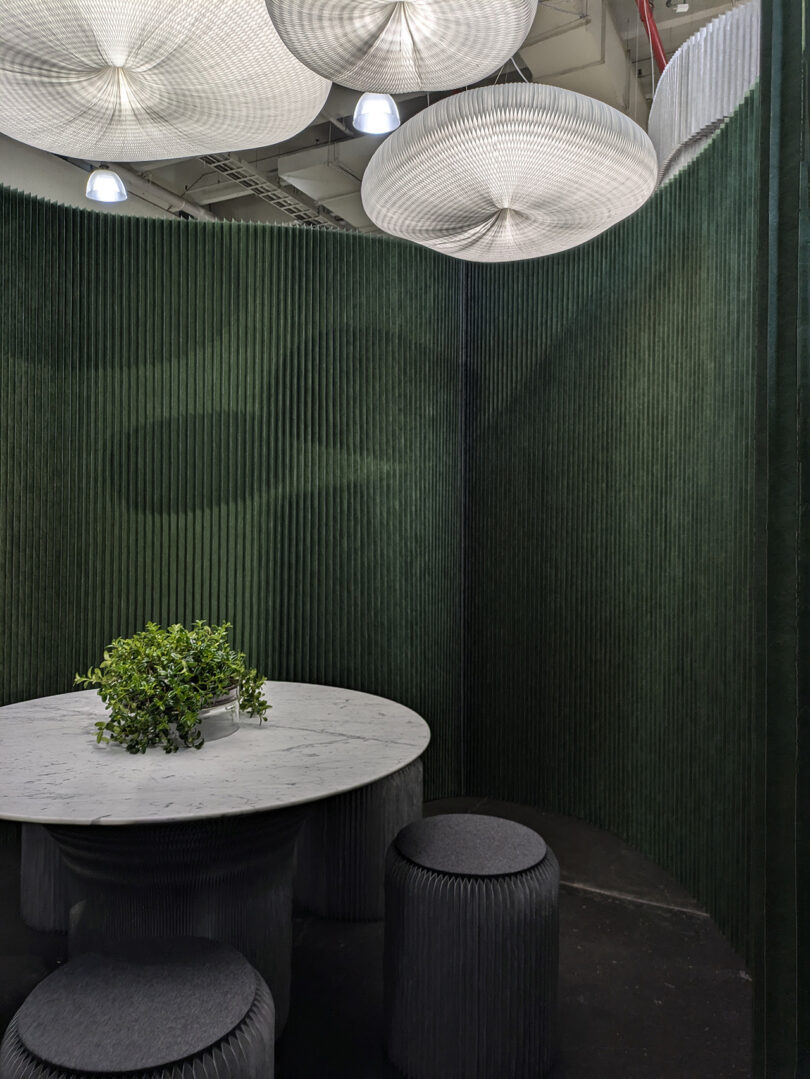 A round marble table with a potted green plant is surrounded by dark, ribbed stools and green textured partitions under large, circular ceiling lights.
