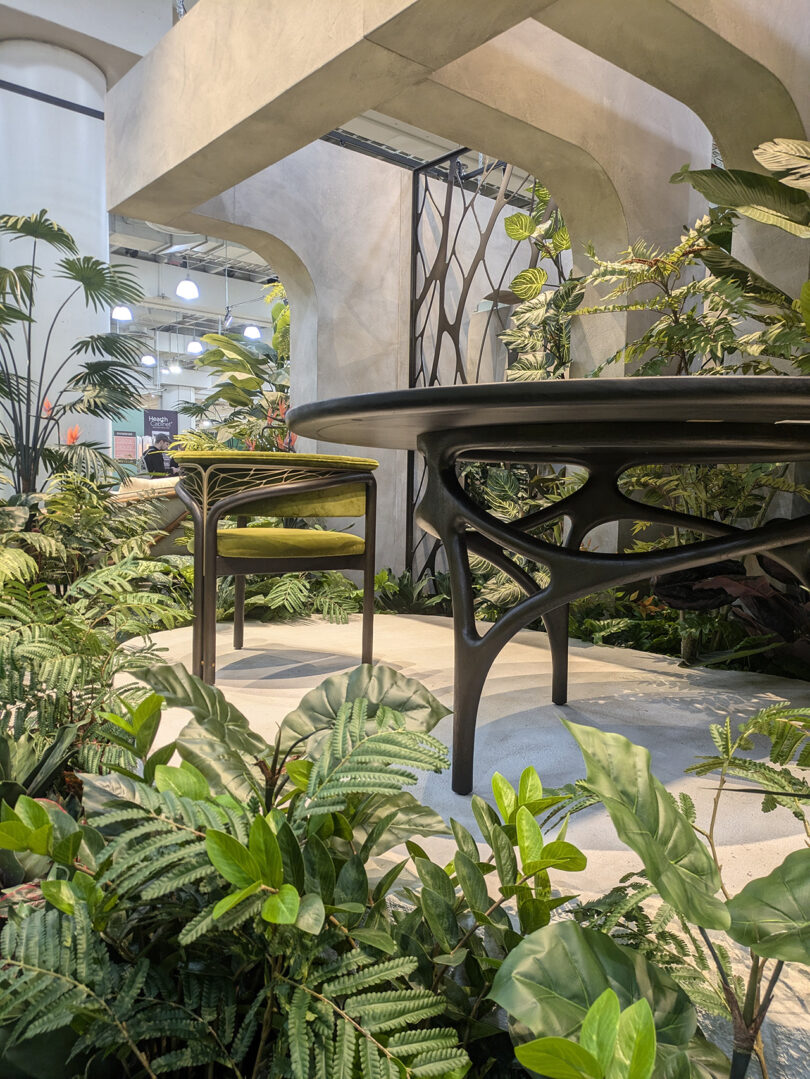 A modern table and chair set is placed on a concrete floor surrounded by lush green plants in a contemporary indoor space with architectural details.