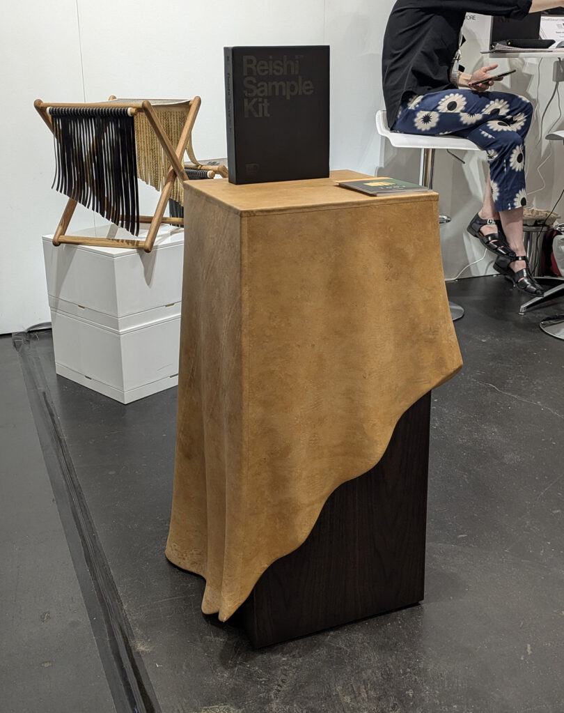 A pedestal draped with a tan, natural-looking material displays a box labeled "Reishi Sample Kit"; a magazine rack and person sitting in the background.