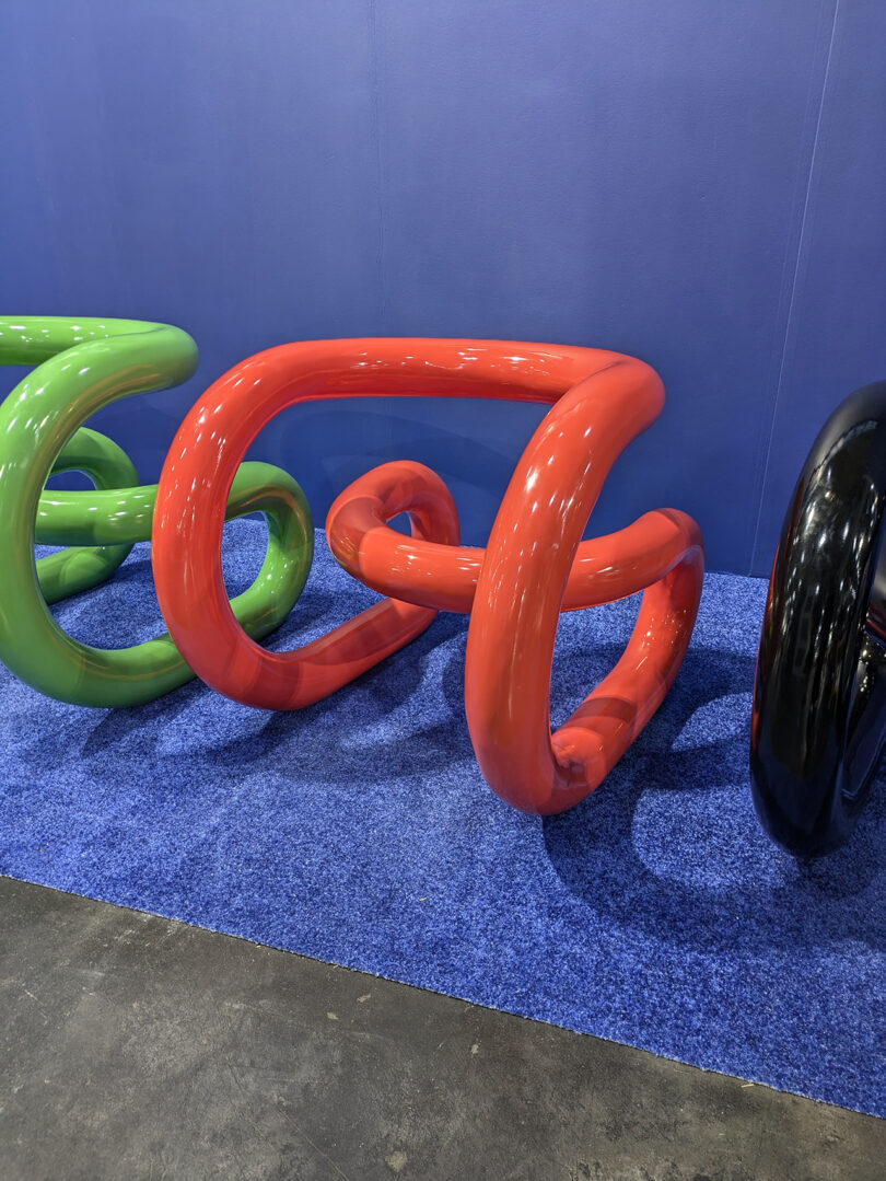 Shiny, abstract sculptures in green, red, and black are displayed on blue carpet against a blue wall.