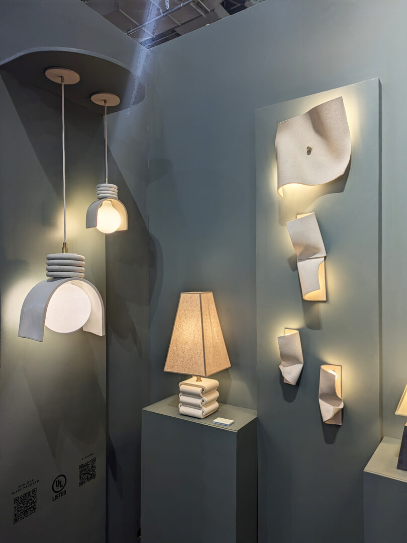 Modern lighting fixtures, including pendant lights, a table lamp, and wall sconces with geometric and abstract designs, displayed against a muted green wall.