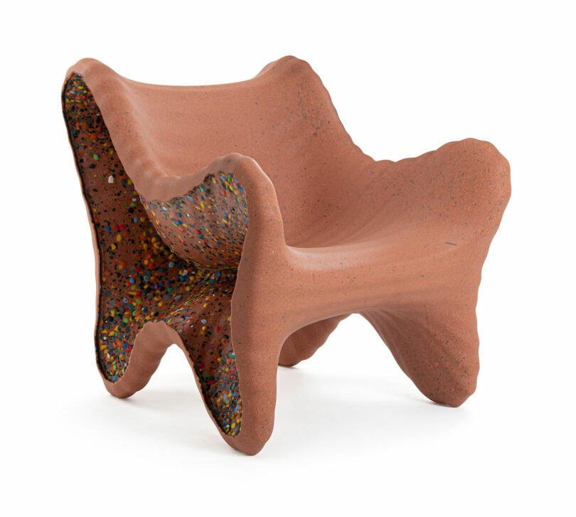A sculptural chair made of reddish material with organic, wavy edges and multicolored speckles on the interior surfaces, reminiscent of In-Side Thomas Heatherwick’s innovative design approach.