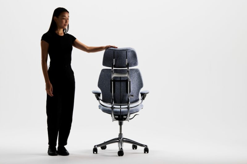 Woman in black standing beside a sculptural ergonomic office chair with a tall backrest and chromed frame on casters.