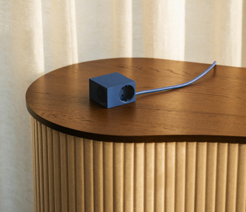 A blue Kord cube-shaped power outlet with two sockets and a blue cord sits on a wooden table in front of a beige curtain.