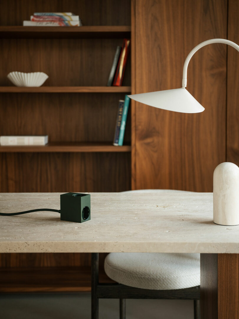 A minimalist desk features a Kord cube-shaped power outlet, a sleek white lamp, and a chair facing wooden bookshelves with select books and decorative pieces.