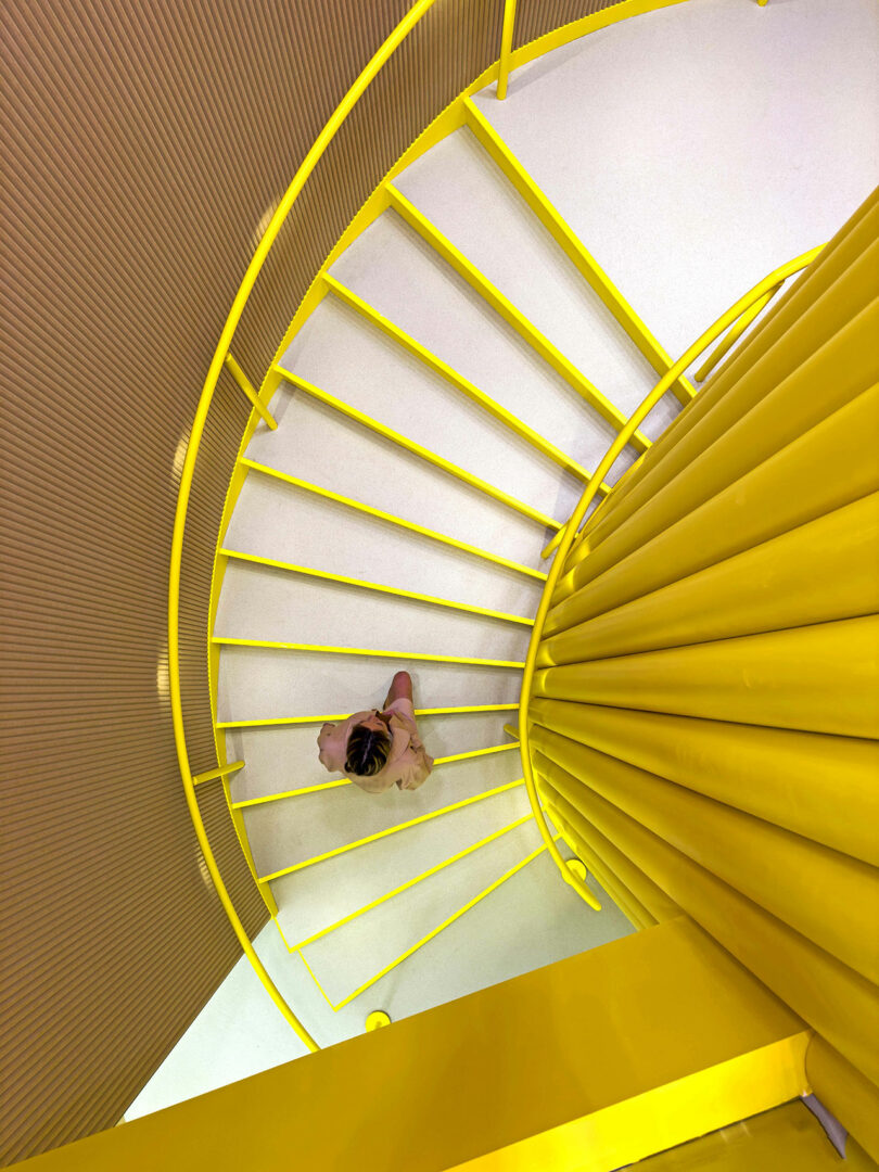 A person descends a yellow spiral staircase, seen from above, with bright yellow railings and a central column dominating the composition—almost resembling a giant LEGO structure in its bold, playful design.