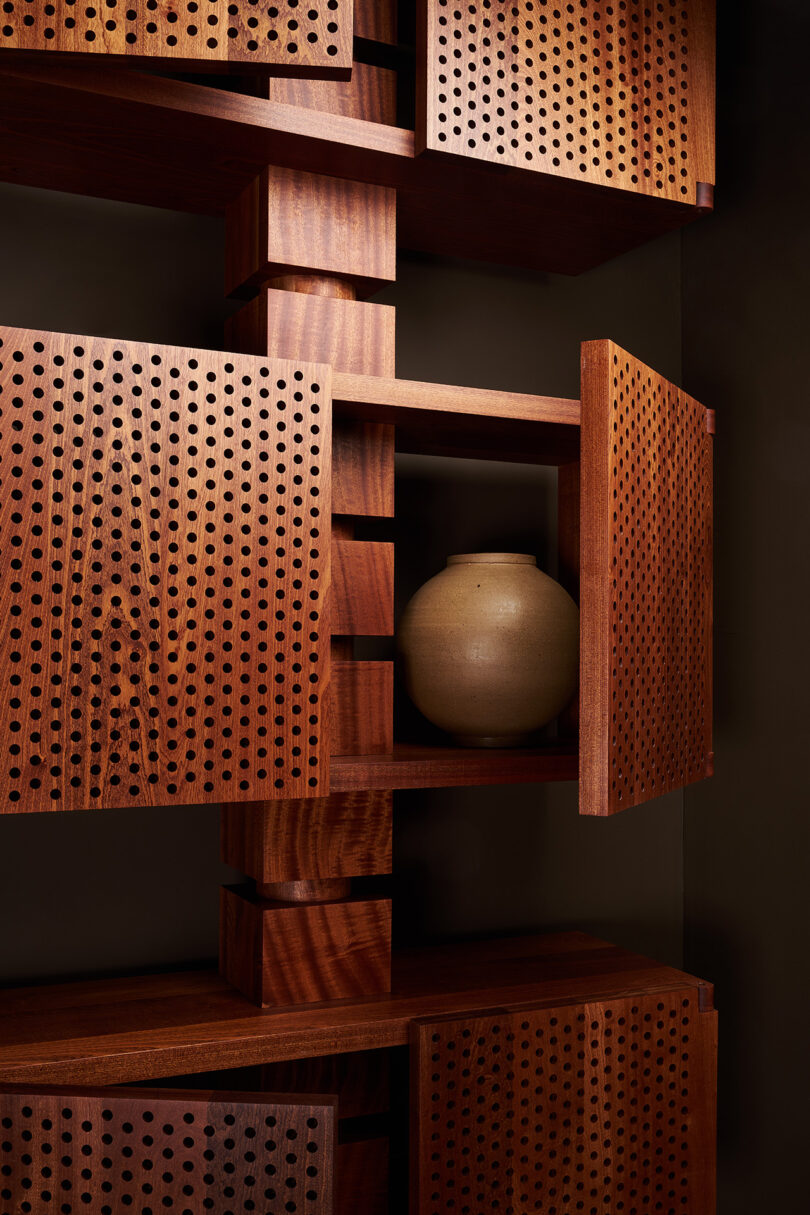 A wooden shelving unit with perforated cabinet doors is partially open, revealing a beige ceramic vase inside.