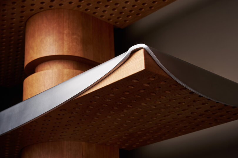 Close-up of a wooden structure with cylindrical and rectangular shapes, featuring a perforated pattern and a thin metal sheet draped over the edge.