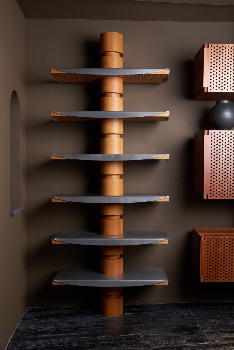 Wood and metal corner shelving unit with six wide, flat shelves mounted to a vertical cylindrical wooden support in a minimalist room with dark walls and floor.