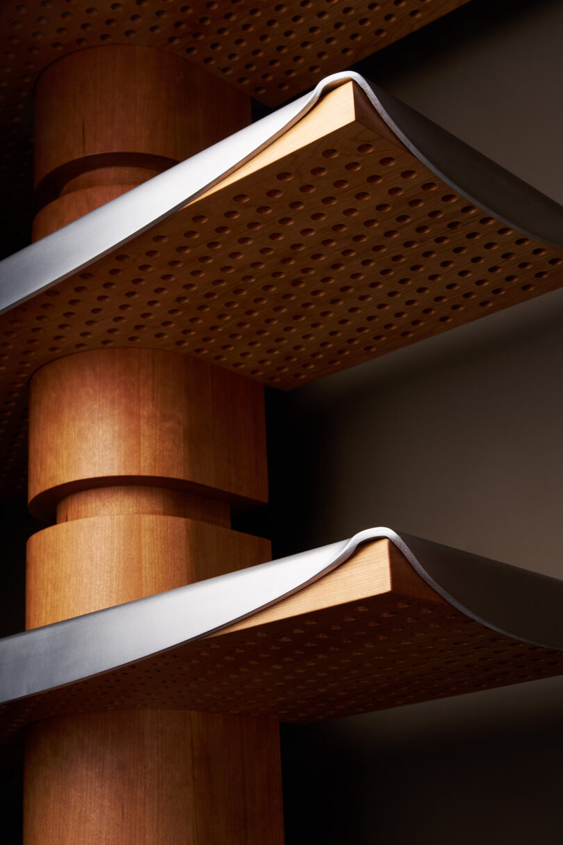 Close-up of two modern wooden shelves with perforated surfaces and metal edges, attached to a cylindrical wooden support.