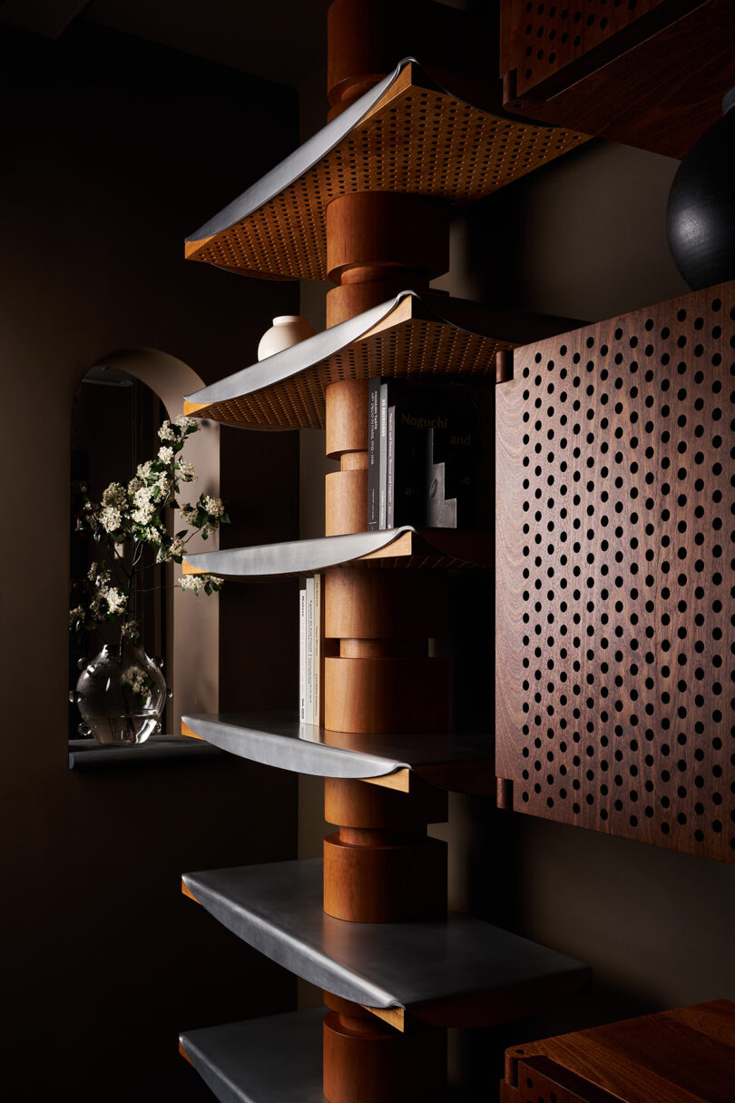 A modern shelving unit with wooden and metal elements stands next to a window with a vase of white flowers and a stack of books on the shelves.
