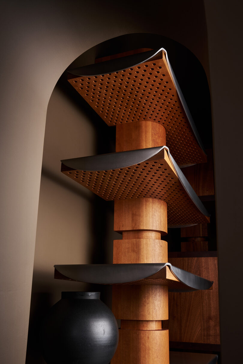 A modern wooden shelf with curved, perforated panels stands against a brown wall under an archway, with a round black vase at its base.
