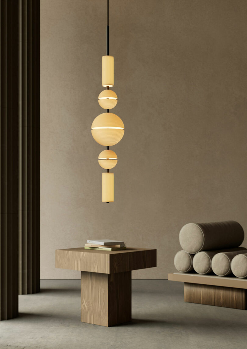 Minimalist interior featuring a Lee Broom geometric pendant light, wooden table with books, and cylindrical cushions on a bench set against beige walls and columns.