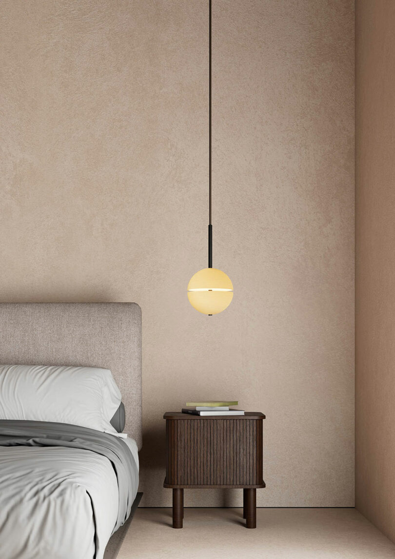 A minimalist bedroom with a beige bed, a small wooden nightstand topped with books, and a Lee Broom round pendant light elegantly hanging above the nightstand.