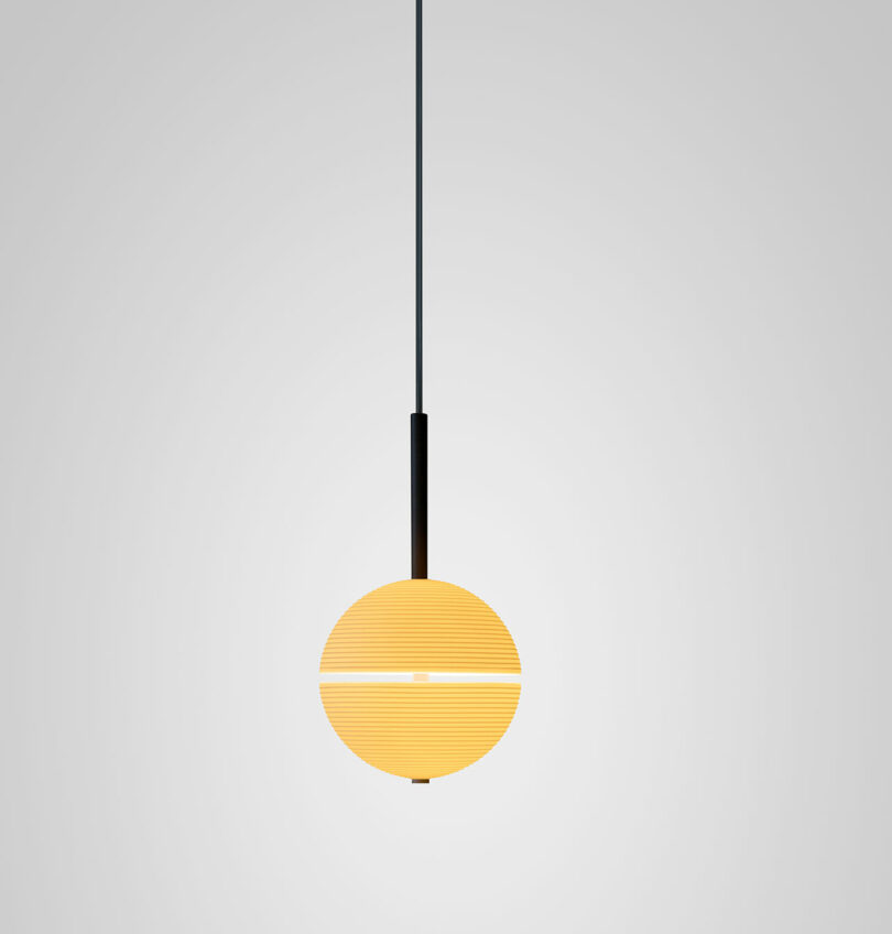 A single modern Lee Broom pendant light with a round, yellowish lampshade hangs from a black cord against a plain, light gray background.