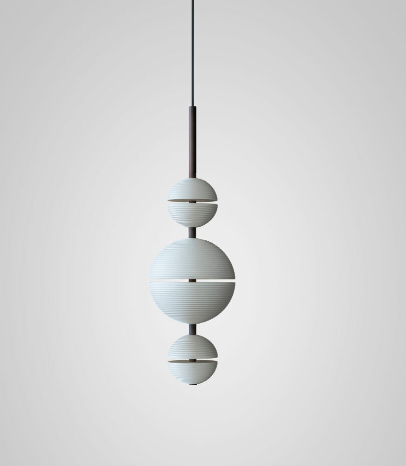 A modern Lee Broom pendant light fixture with three stacked, white, ribbed spherical shades hanging from a black cord against a plain light gray background.
