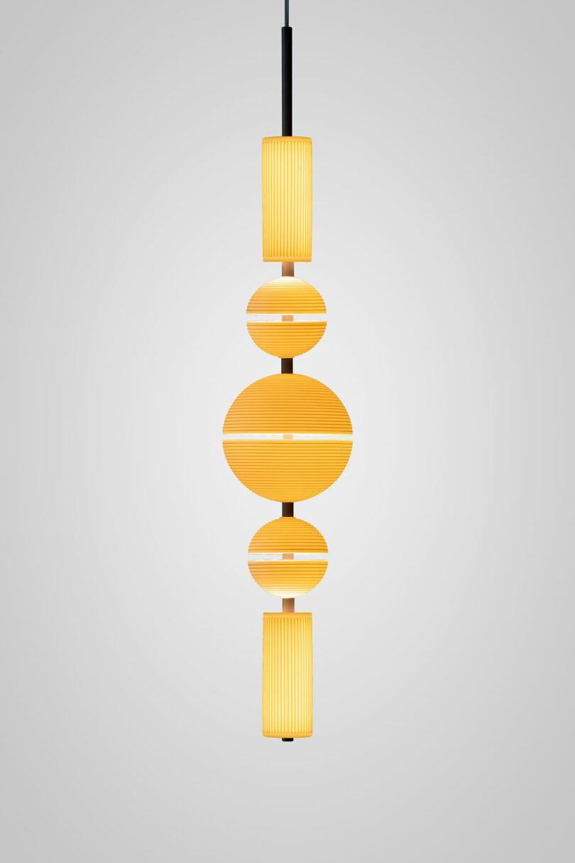 A modern Lee Broom pendant light features geometric, ribbed yellow and white shades arranged vertically on a black cord against a plain gray background.