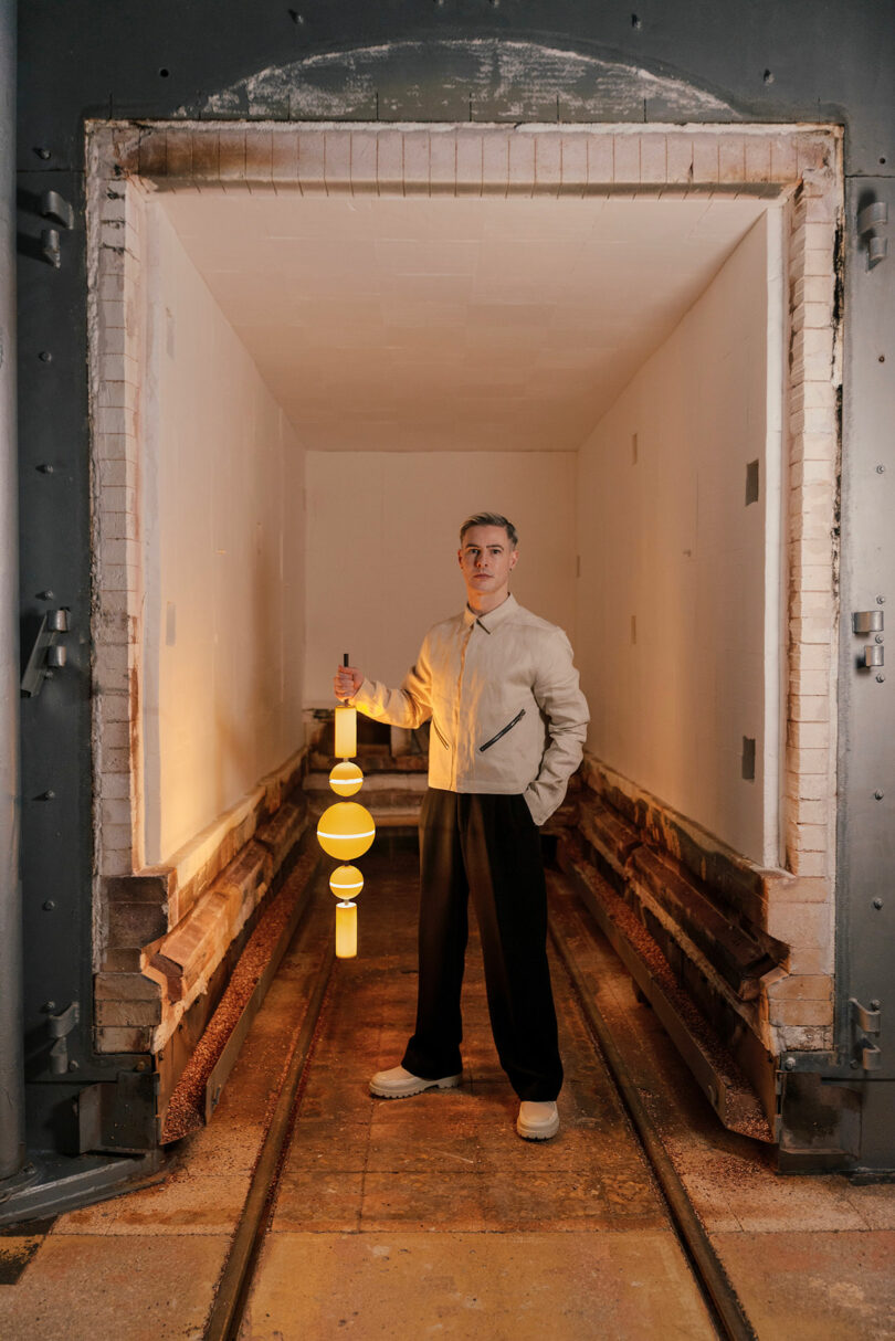 A person stands in an empty industrial chamber, holding a vertical, glowing yellow Lee Broom light fixture with circular elements.