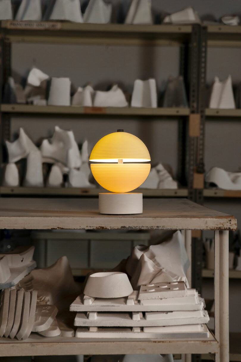 A spherical yellow Lee Broom lamp sits on a table in front of shelves filled with white plaster molds in a workshop.