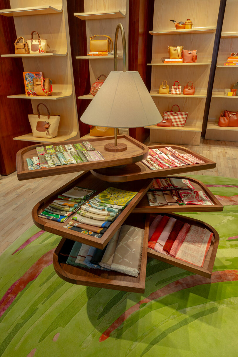 A tiered wooden display with trays of folded scarves surrounds a lamp; shelves of handbags are visible in the background.