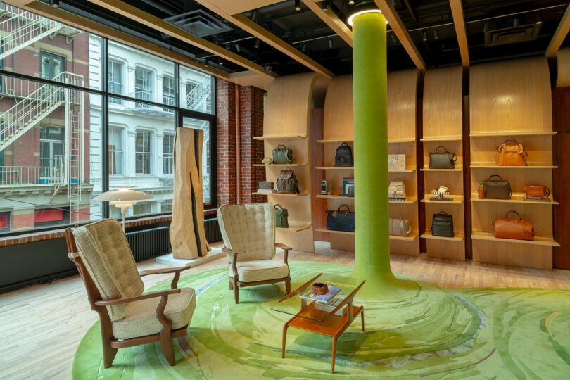 A modern boutique interior with two armchairs, a small table, green carpet, a central green column, shelves displaying bags, and large windows with a city view.