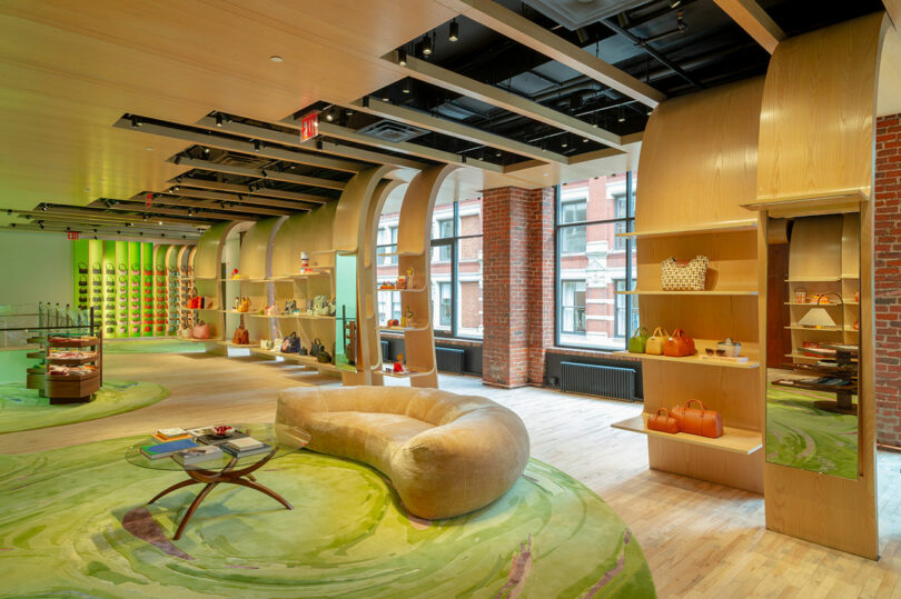 Modern retail store interior with curved wooden shelves, display tables, green patterned carpet, large windows, and various handbags and accessories on display.